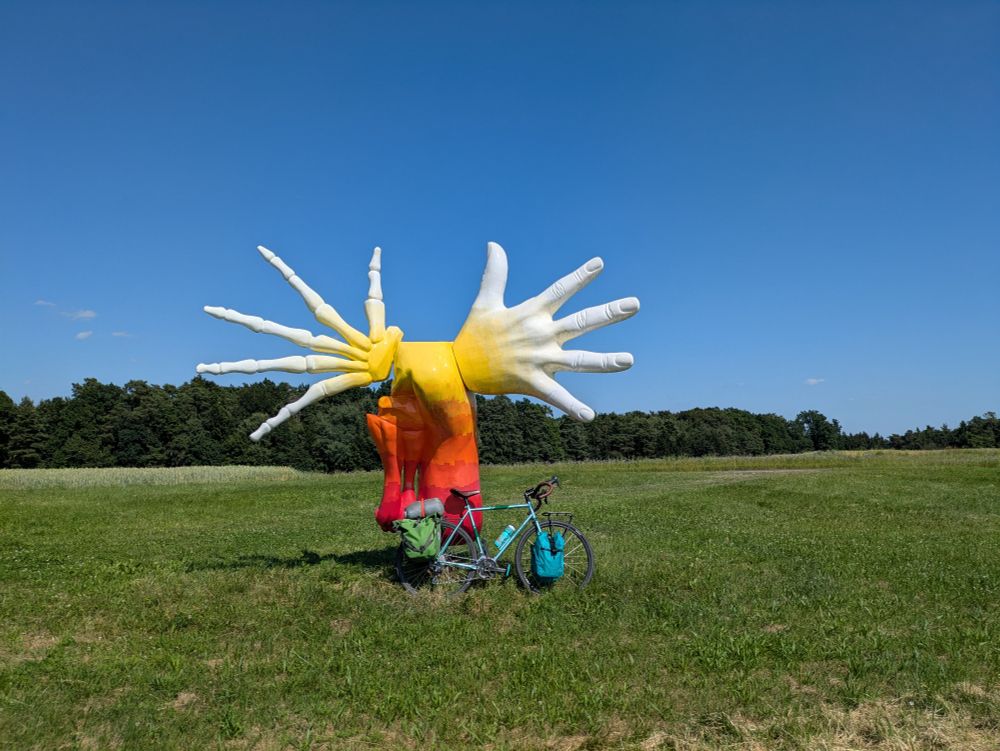Bike in field under sculpture of fleshy hand and bony hand.