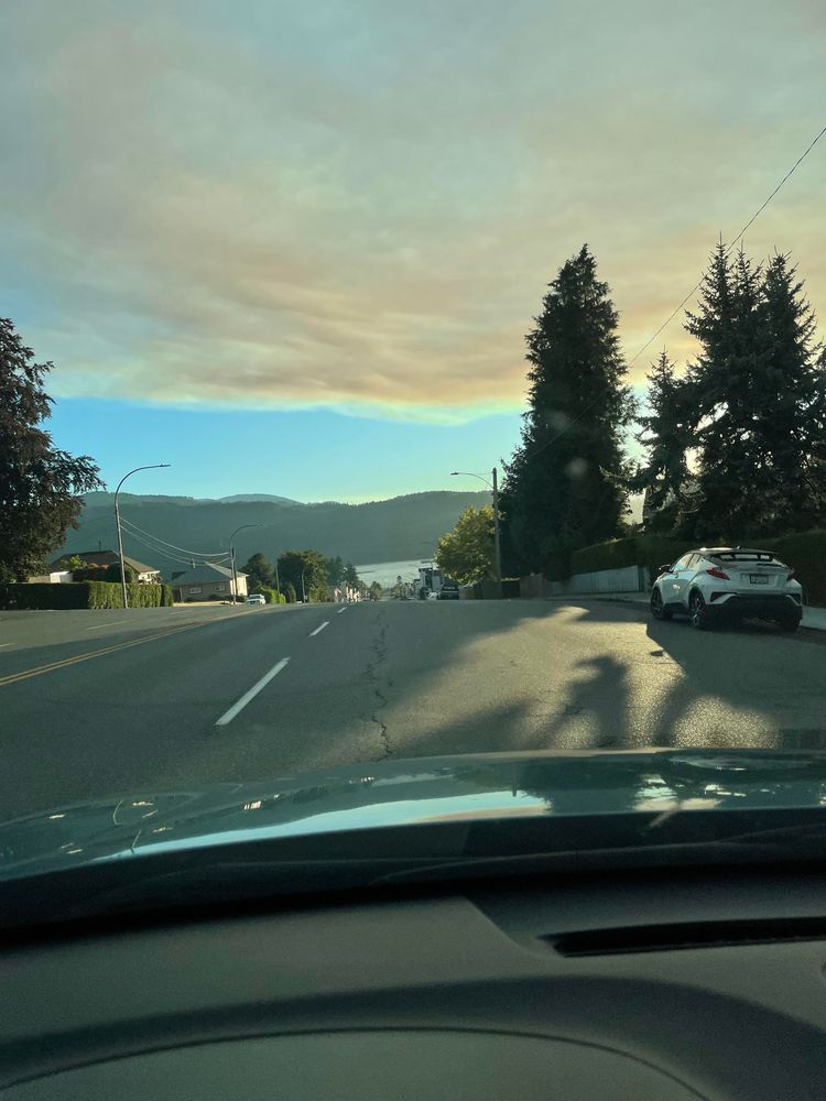 A blanket of smoke is encroaching overtop the city. There is a band of blue sky to the west where the sun is going down behind the trees. The picture is out the dash of a car driving on a wide empty 4 lane street