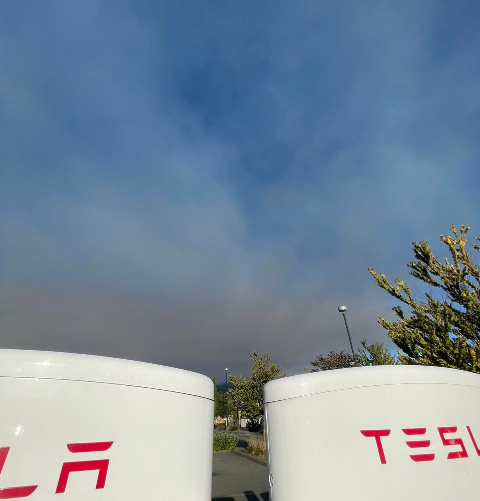 A pall of smoke looms over a ridge in the background behind a bank of tesla chargers