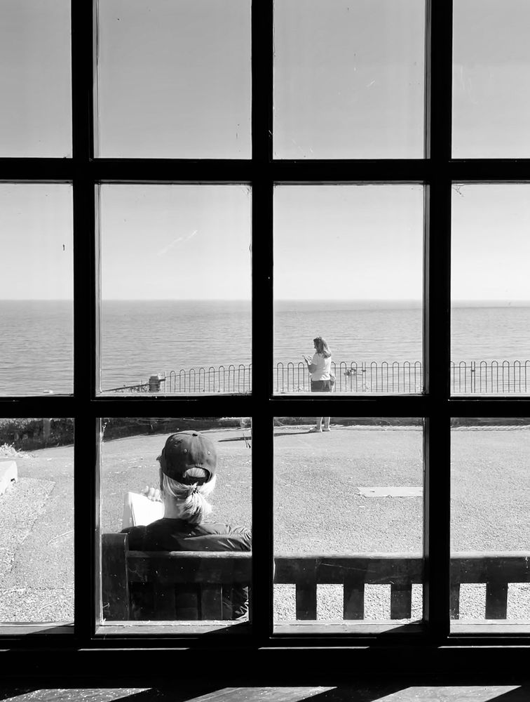 Black and white view from behind a framed window of a woman sitting on a bench reading. Another woman is standing looking out to sea. 