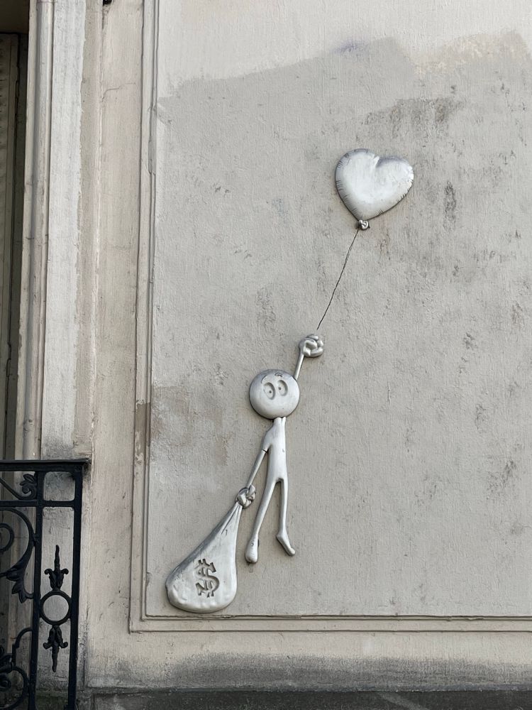 A piece of street art affixed to a grey wall in a metallic medium. A childlike image of a person holds a helium balloon in their left hand. The balloon is carrying them to the sky. In their left hand is a sack with a dollar sign motif on it. The weight of the sack is keeping the person trapped on the ground. 