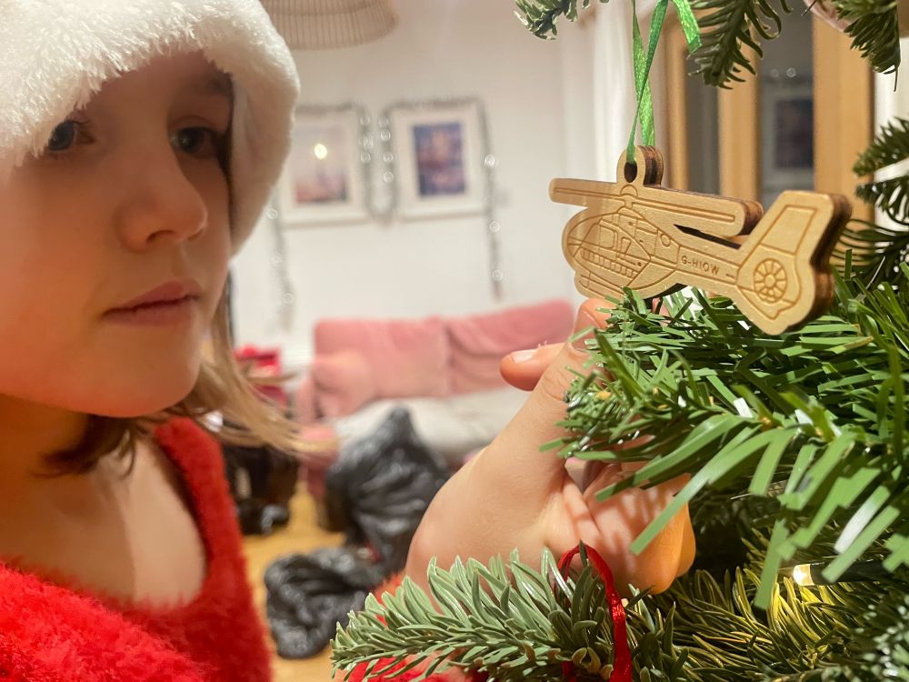 Child in festive hat holds helicopter shaped wooden decoration. The decoration is hanging from a Christmas tree and is engraved with the letters G-HIOW. 