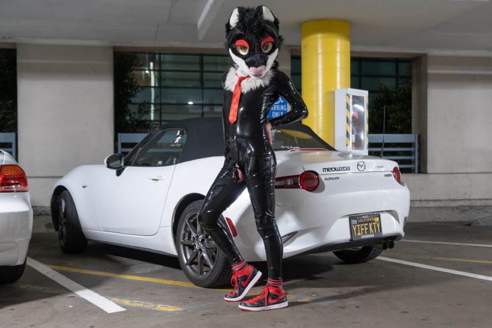 Jamie in latex standing in front of a white MX-5 Miata with a license plate that reads YIFFKTY