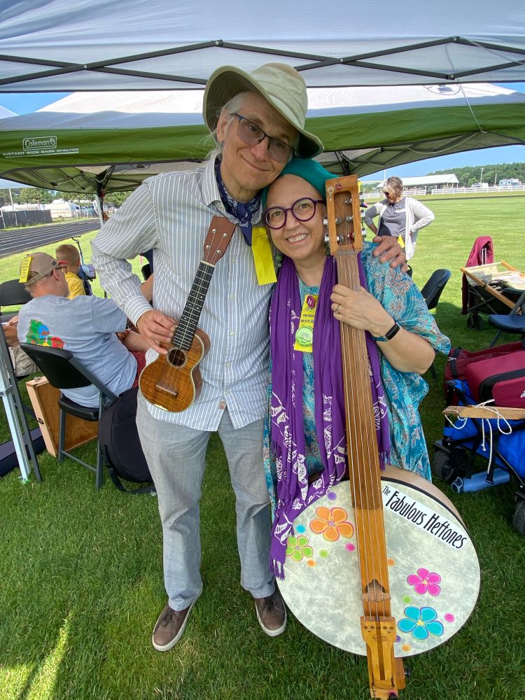 Two musicians, Brian Hefferan and Lynn Hershberger Hefferan (The Fabulous Heftones) outside on a sunny morning at a music festival. They are wearing ribbons and buttons that identify them as instructors. Brian is 5‘10“ and white haired in his young 60’s. He is wearing a summer fabric hat, white pinstriped dress shirt untucked, with khaki pants and brown casual shoes. Lynn wears a turquoise caftan she sewed from African wax print Fabric and a purple sarong as a scarf around her neck. She is wearing a wool beret.  Both are smiling and wear glasses. Brian is holding a soprano ukulele, a small instrument with four strings that looks something like a shrunken guitar. Lynn is holding a Heftone bass which looks like a huge banjo with retro 70’s flower stickers on the front. The hand-carved wood neck of the bass is almost as tall as Lynn, who is 5’2”. The bass has five strings-EADGC