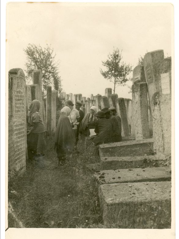 A black and white photo of a group of people gathered in a cemetery. Three women, professional mourning women, wearing shawls on their heads, one of them leads prayers from a prayer book 