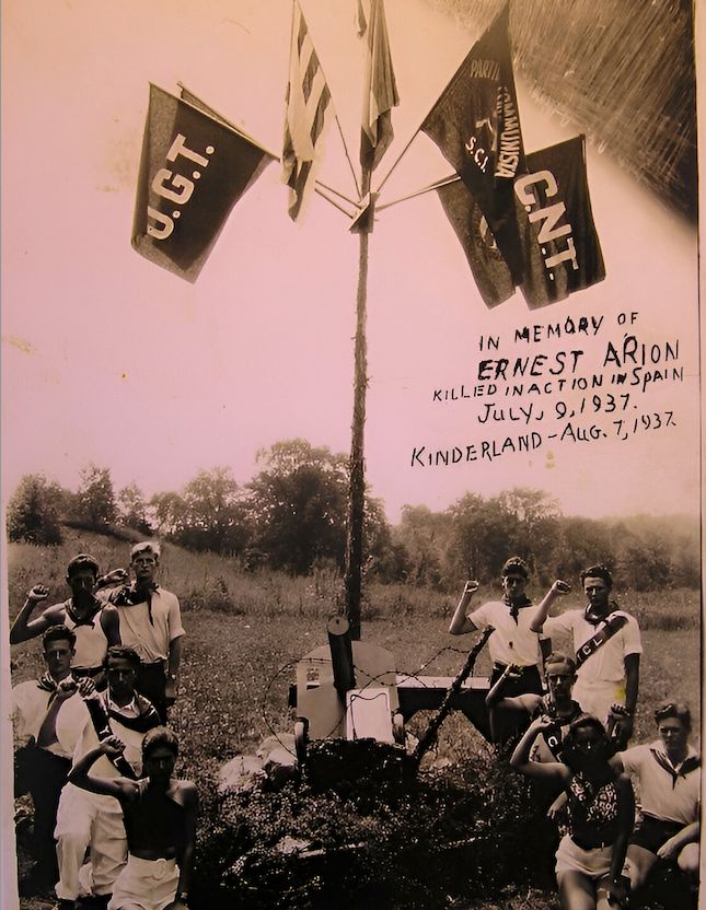 A group of young people pose, fists raised, around a makeshift monument to their fallen comrade Ernest Arion who died fighting in the International Brigades 