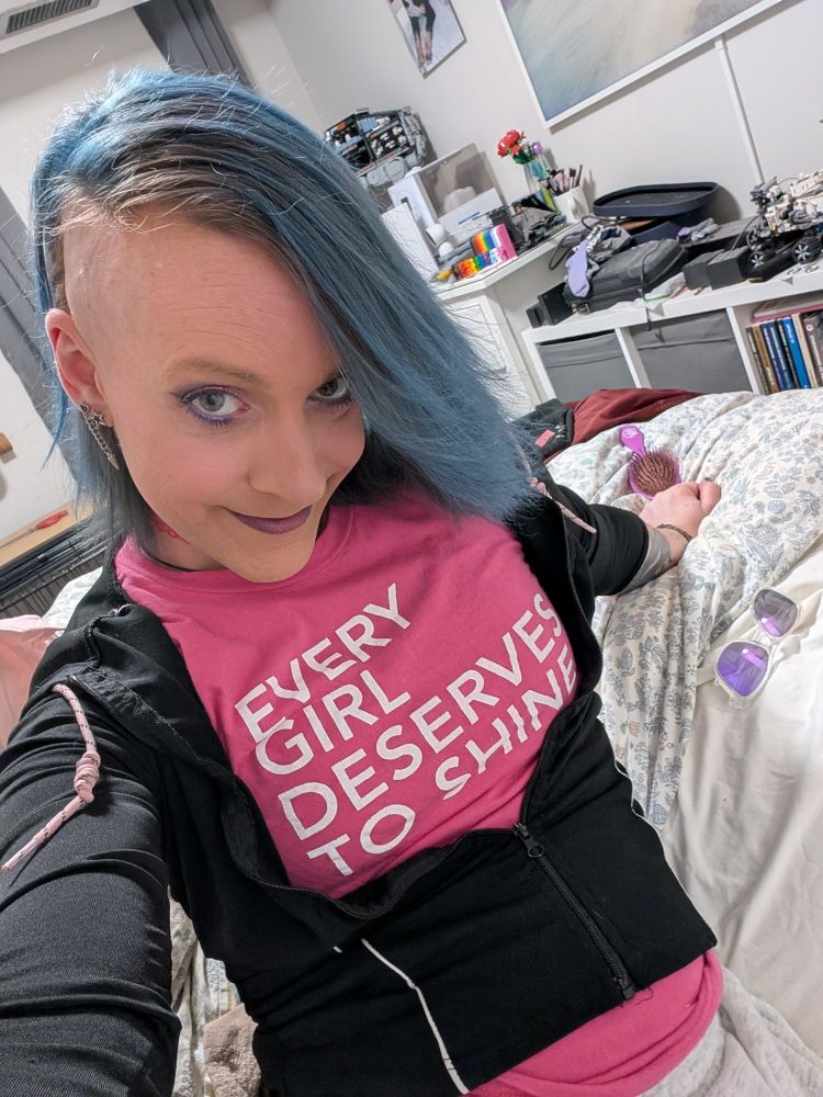 kaylee taking a selfie, sitting on her bed, wearing a black hoodie over a pink t-shirt that reads "EVERY GIRL DESERVES TO SHINE", her hair is dyed blue, straightened and shaved on one side.