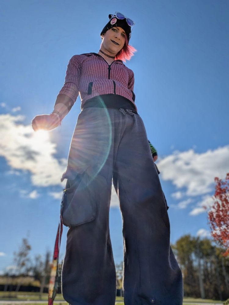 Kaylee outside, standing, seen from a low angle and backlit by the sun, wearing her toque with kitty ears, purple sunglasses, dark makeup, a pink hoodie and grey sweatpants