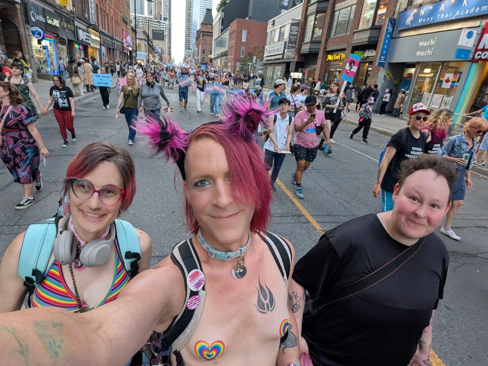 Kaylee, Jessica and Emmett walking in a crowd in the middle of Yonge St in downtown Toronto 

Kaylee has her top off and rainbow-colored, heart-shaped pasties over her nipples