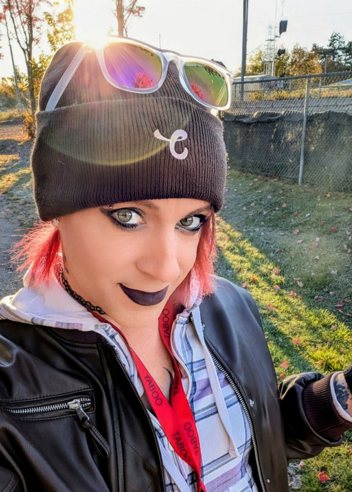 kaylee outside, backlit by the sun, smiling and standing in the grass in front of a chain-link fence, a rail line and some trees. she's wearing dark makeup, a brown jacket, white & purple plaid hoodie, a taboo show lanyard, a brown chaturbate toque and purple sunglasses on top of her head. her hair is shoulder-length, straightened and dyed red.