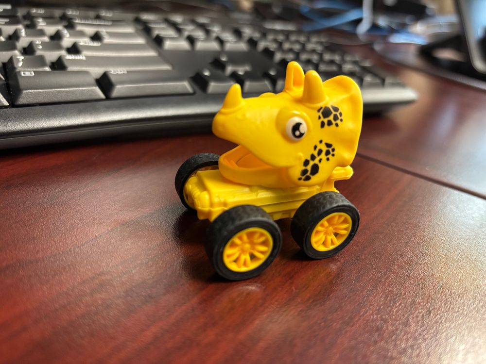 A yellow toy care with the head of a triceratops sits in front of a keyboard on a desk.