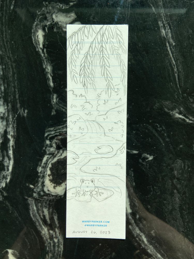 Image shows a plain white bookmark that has been sketched on. The sketch is of a tranquil pond scene, with weeping willow branches hanging from the top of the bookmark over a pond with lily pads. The frontmost lily pad has a cartoon frog sitting on it.