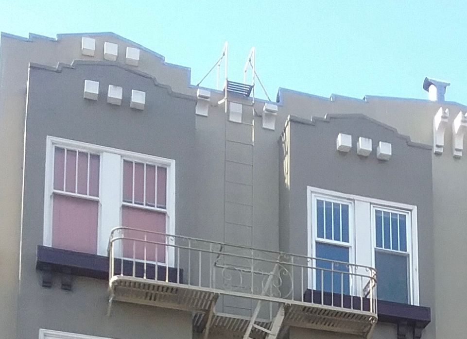 Street side view of four upper story windows on a grey painted apartment building—two with pink window shades, and two with blue window shades.