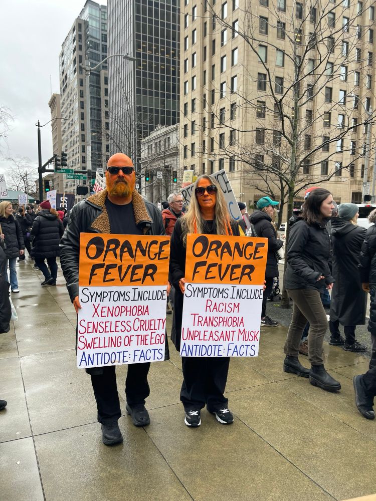 Two protesters painted orange holding protest signs with the symptoms of Protest Fever