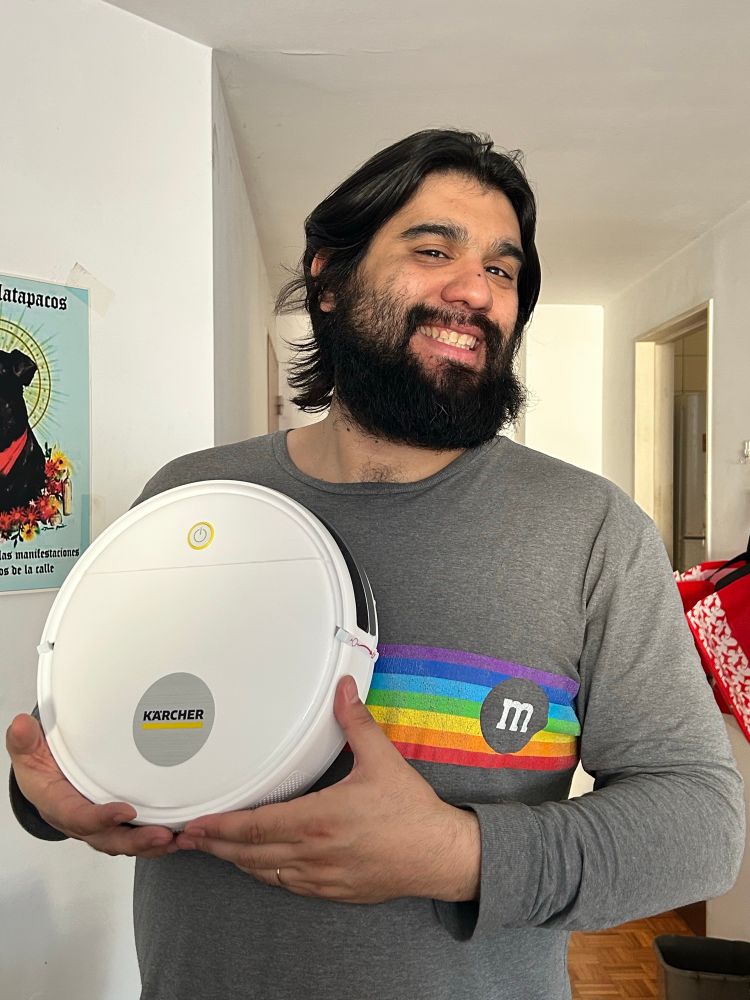 Un hombre muy peludo posando con un robot aspiradora