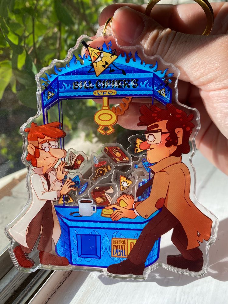 A keychain featuring artwork of Bill Cipher, Fiddleford McGuckett and Stanford Pines the characters are depicted playing at an arcade claw machine.