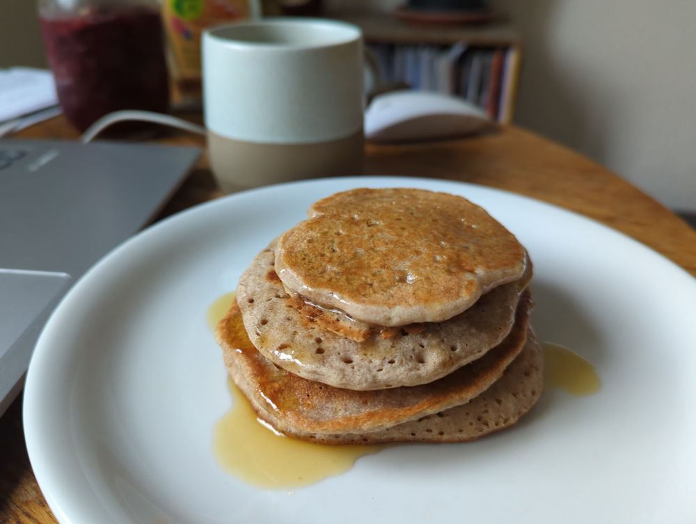 Ein Stapel Pancakes, die mit Ahornsirup übergossen sind, auf einem weissen Teller. Hinten eine Kaffeetasse, links ein grauer Laptop.
