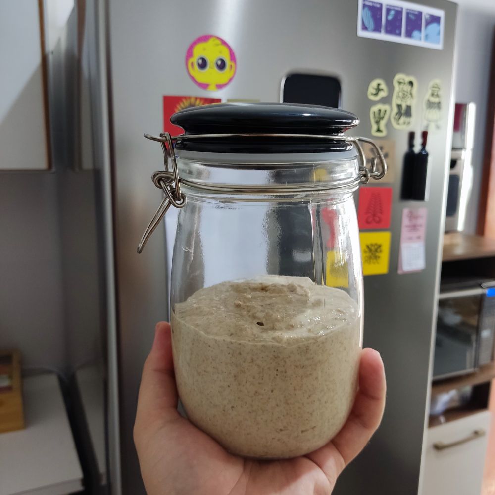 Um pote de vidro com fermento natural até a metade. É uma massa úmida com textura que parece areia encharcada. Pelo menos visualmente.