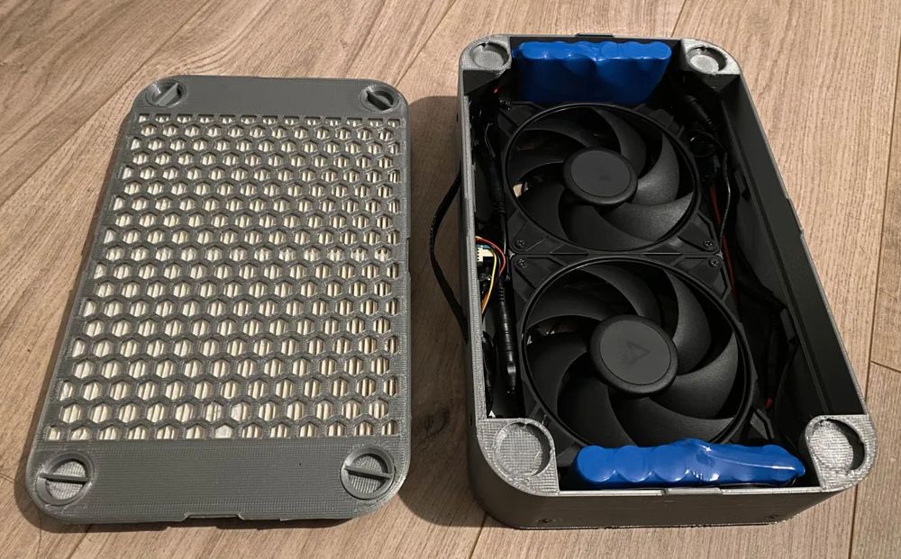 Photo d'un purificateur d'air portatif type Koala ouvert par son coté de la grille-filtre. Les deux ventilateurs noirs sont visibles ainsi que les deux batteries bleues et divers câbles reliant le tout.

L'appareil est posé sur le sol.