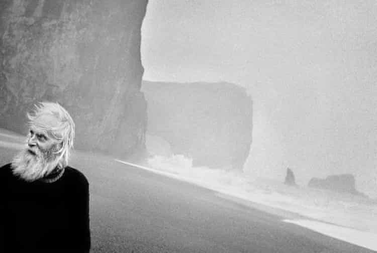 Un uomo con barba e capelli bianchi cammina sulla spiaggia islandese. 