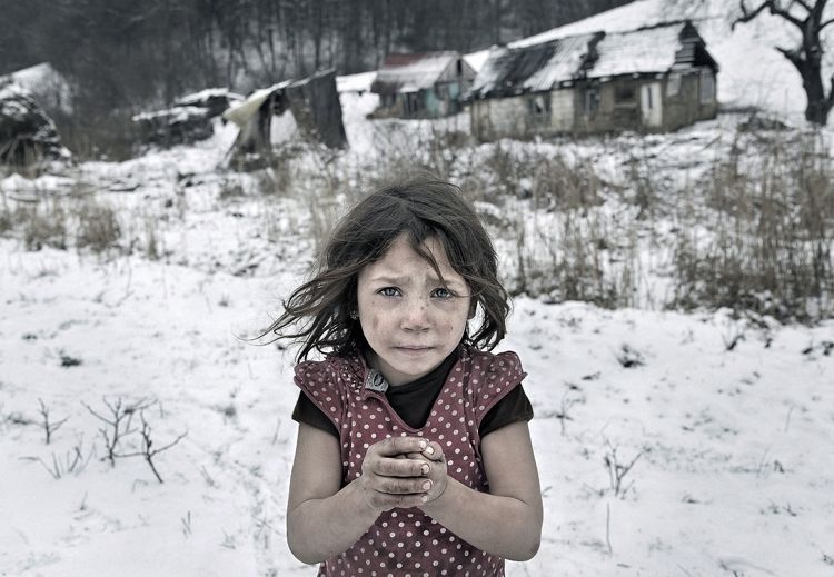 Una bambina in abiti leggeri sta in piedi nella neve. Sullo sfondo si intravedono delle baracche di legno.