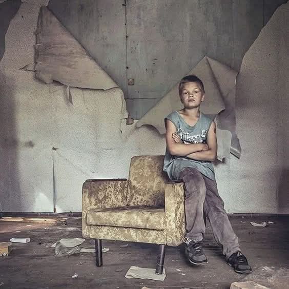 Un ragazzino, seduto sul bracciolo di una poltrona, in una stanza spoglia e fatiscente. 