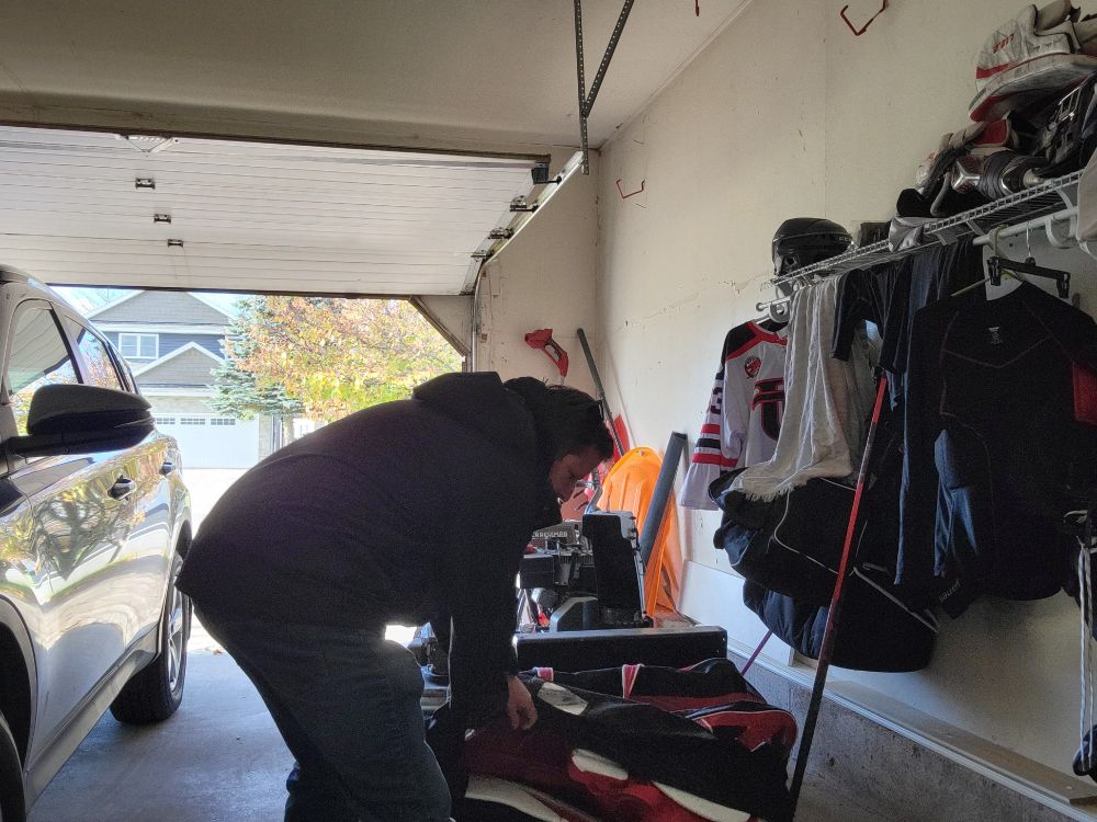 A person packing a hockey bag in a garage. A wall of gear is visible to the side