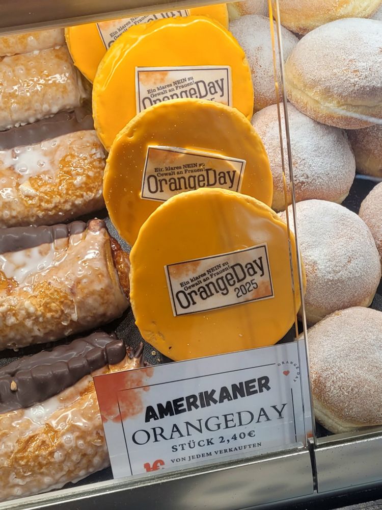 Das Bild zeigt eine Auslage in einer Bäckerei oder einem Café. Zu sehen sind verschiedene Backwaren, darunter Amerikaner mit Zuckerguss und Schokoladenüberzug, sowie Berliner mit Puderzucker. Besonders hervorgehoben werden jedoch die "Orangeday"-Amerikaner, die mit einer orangenen Glasur überzogen und mit einem Aufkleber versehen sind, der auf den "Orangeday" hinweist und eine klare Ablehnung von Gewalt gegen Frauen zum Ausdruck bringt. Das Preisschild gibt an, dass ein Stück 2,40€ kostet und ein Teil des Erlöses gespendet wird.