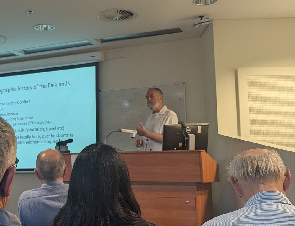 Dave Briton, an affable, bearded white guy in a white linen shirt, standing at a lecturn with a PowerPoint presentation behind him.