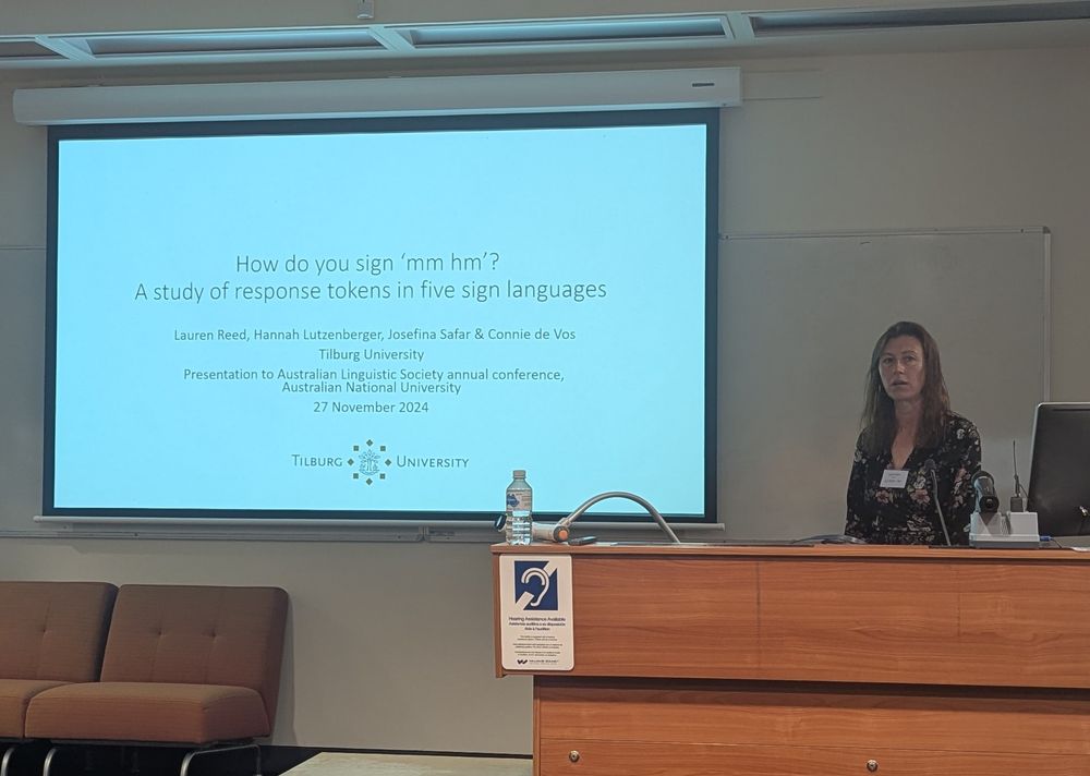 Lauren Reed, a white woman with a dark dress, stressing at the front of a lecture theatre in front of a white PowerPoint slide with black text