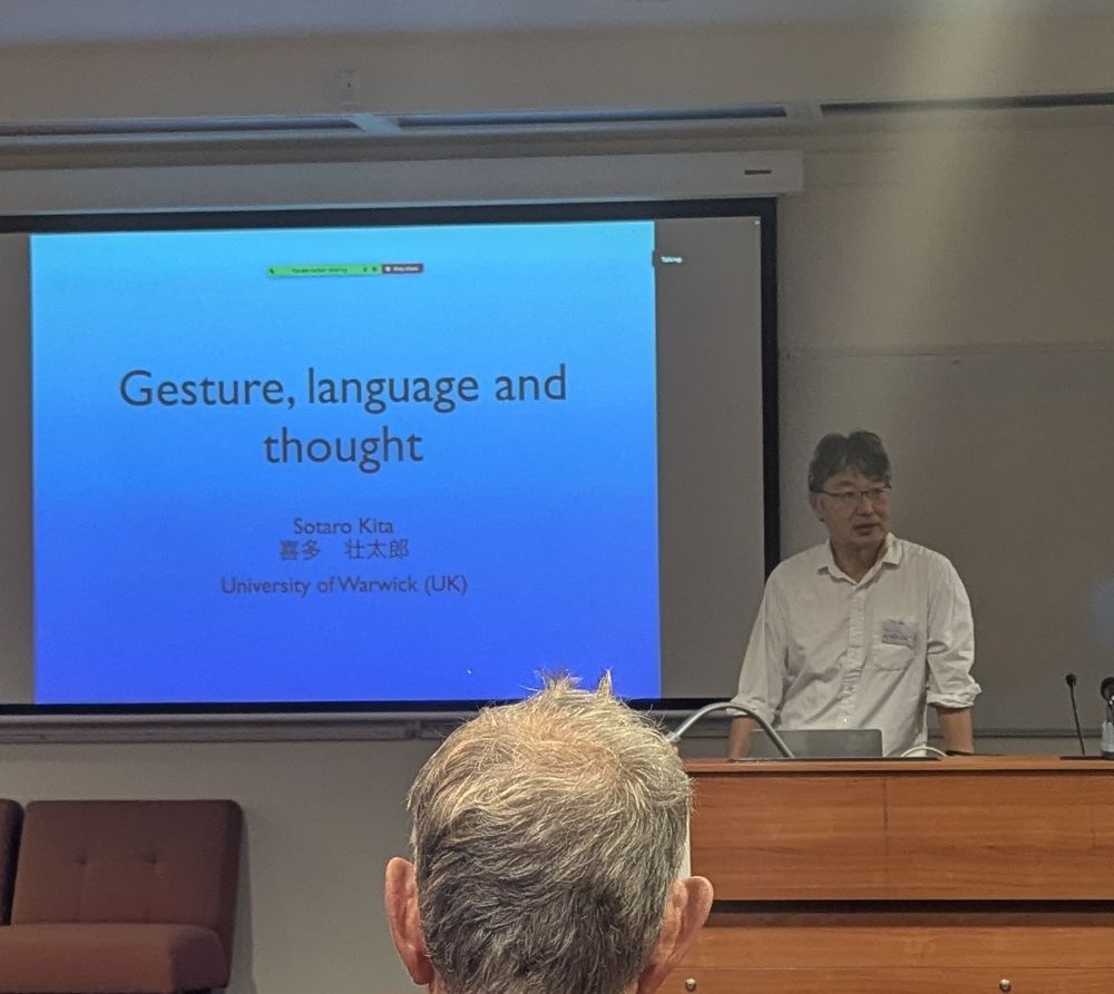 Kita, a Japanese man in a white business shirt with the sleeves rolled up, standing in front of a PowerPoint slide with blue gradient background with black text saying Gesture, Language and Thought.