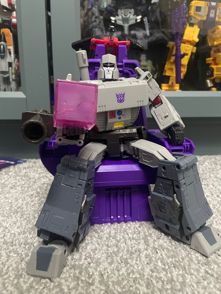 Studio Series 86 Megatron, a light and dark grey robot action figure, sitting on a purple throne. He has a very dark brown cannon affixed to his right arm and is holding a pink Energon cube in the right hand. A red and black robot bird (Laserbeak) is perched on the throne.