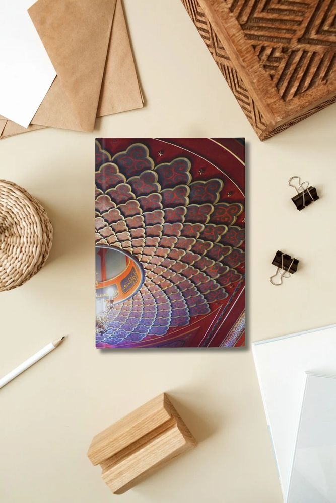 dark red hardcover journal on a warm toned desk. the cover of the journal is a photograph of the roof of a little castle in Trieste, northern Italy. the roof has an intricate decor dark gold and deep red.