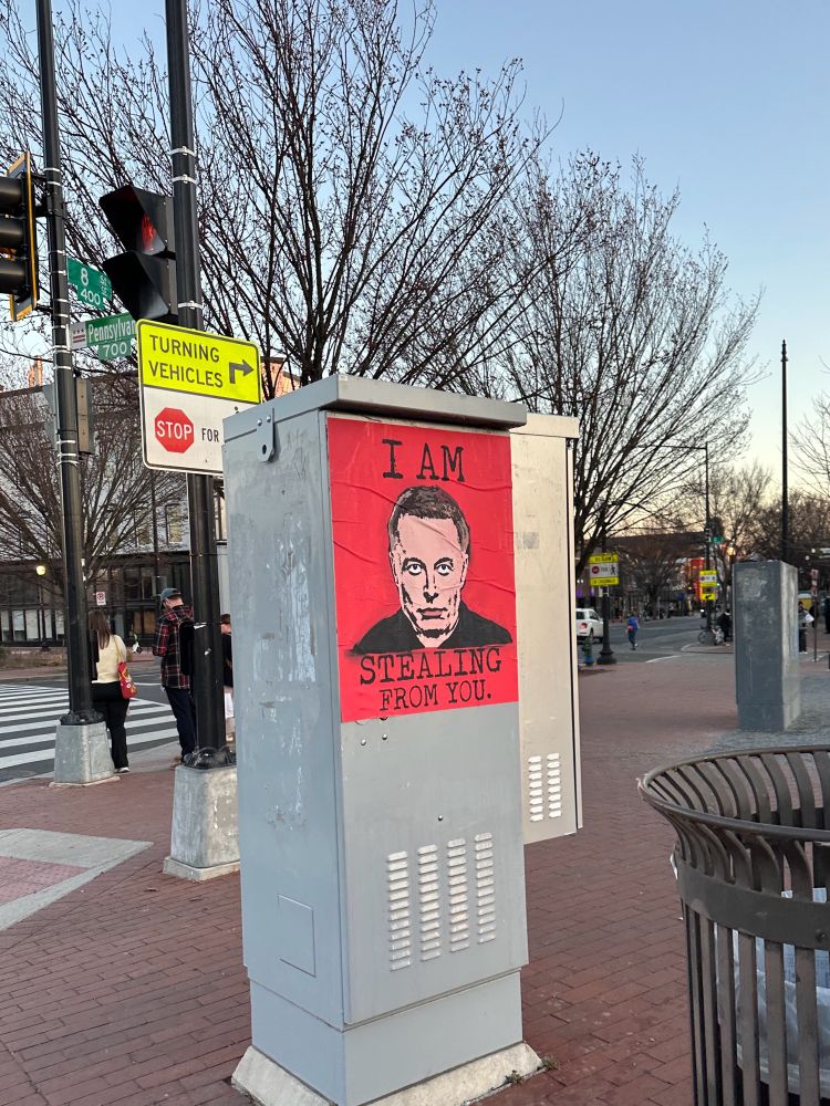 A sign with Elon Musk’s creepy face reads “I Am Stealing From You.”
