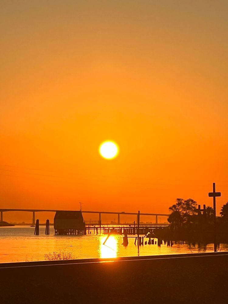 the calm water reflects the orange sun, creating a shimmering path of light. the antioch bridge is in the distance. on the right, a dark silhoue of trees on the shoreline. a small boathouse and multiple pillars stretch across the delta…