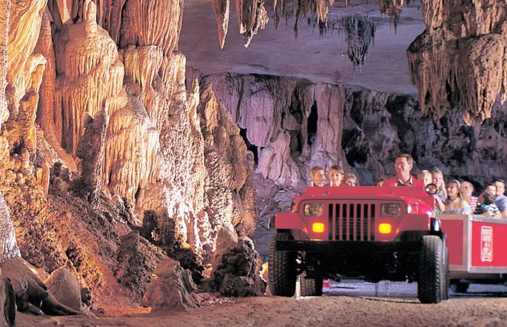 A red open-top cave tour vehicle drives through a stunning underground cavern with passengers inside, surrounded by glowing stalactites and textured rock walls. #TheMotherRoad #Caves #touristattraction