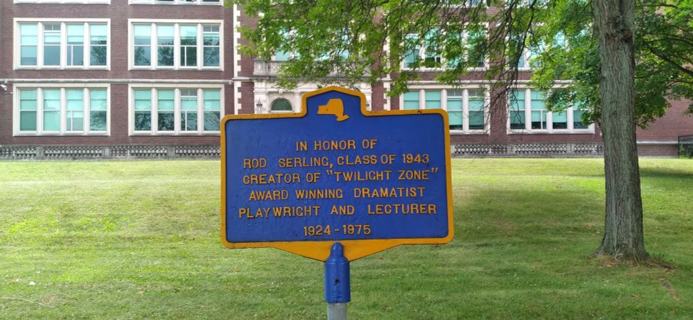 Blue and gold New York state historical marker about Rod Serling, an alum of Binghamton High School, visible in the background at the end of the lawn.