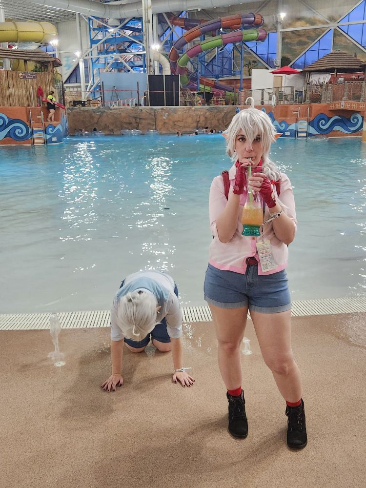 A cosplay of Alisaie and Alphinaud from FFXIV. Alisaie is drinking a drink while Alphinaud is on his hands and knees like he just crawled out of the water after almost drowning.