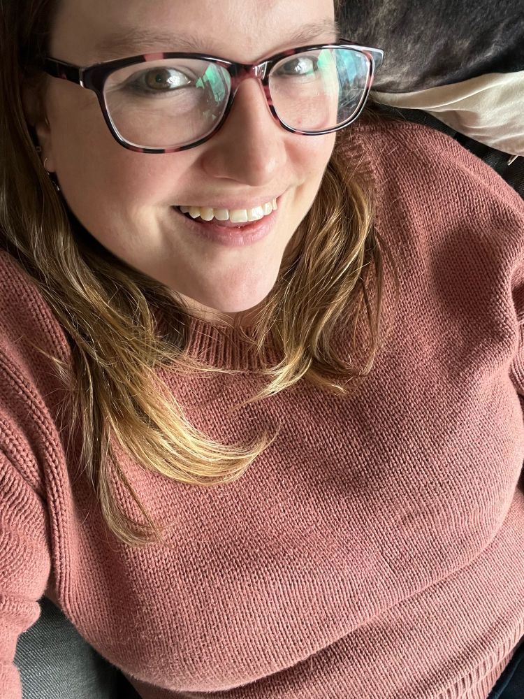 White woman with glasses and medium length light brown hair in a peach sweater is smiling.