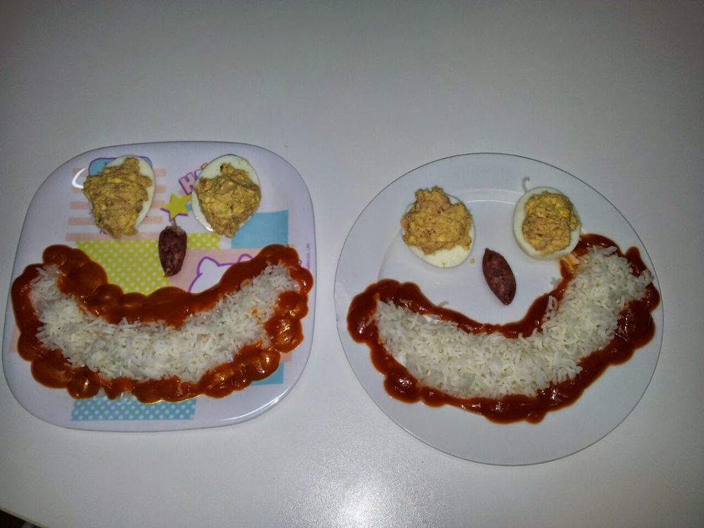 Yo voy a describir lo que tengo delante pero pensad bien si queréis leerlo. Dos platos en los que hay: dos mitades de huevo duro rellenas de atún y mayonesa colocados como si fueran los ojos; una oliva que forma la nariz, y una sonrisa de arroz bordeada con tomate (que serían los labios). Pido perdón de nuevo.
