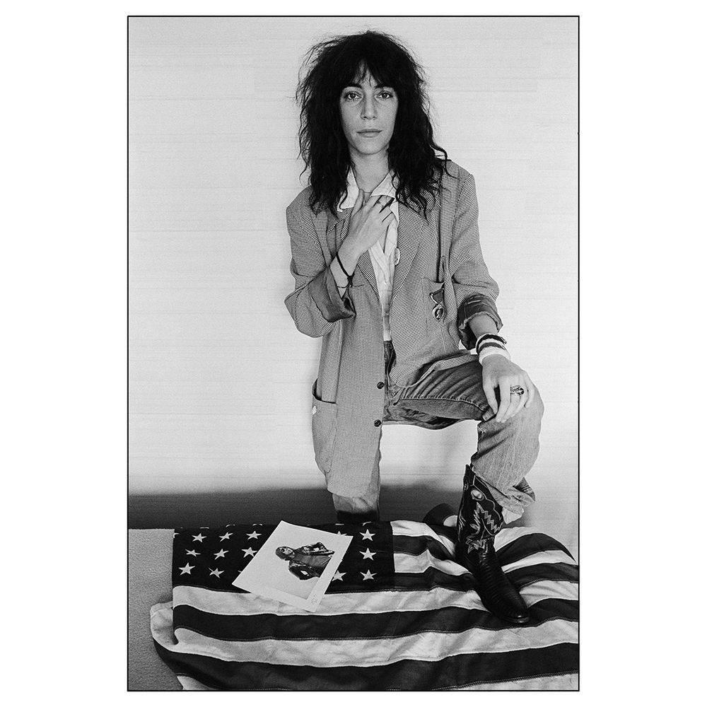 A woman stands facing the camera, her left leg propped on an American flag laid flat on a table. Also on top of the flag is a photograph print of a portrait of a man.