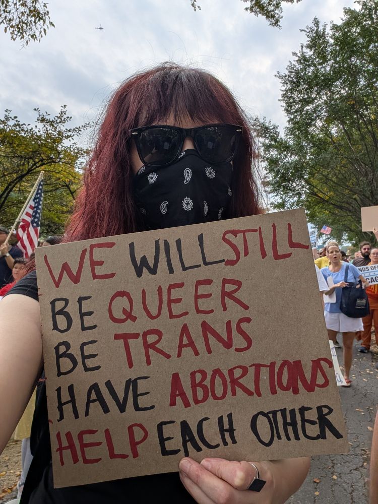 Me in a black tee, black sunglasses, & black paisley bandana over my face, holding a sign reading 
WE WILL STILL
BE QUEER
BE TRANS 
HAVE ABORTIONS
HELP EACH OTHER 