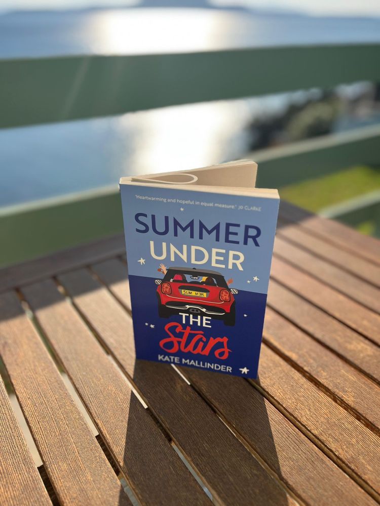 The book “Summer Under the Stars” sits on a wooden table in front of a sunny Greek landscape.