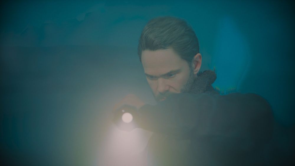 A screenshot of Tim Breaker from the game Alan Wake 2, wielding a flashlight pointed at the camera.