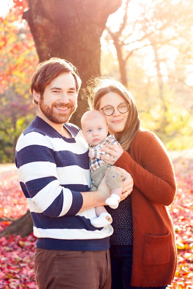 A family portrait of a man and a woman holding a baby in front of a fall background. 