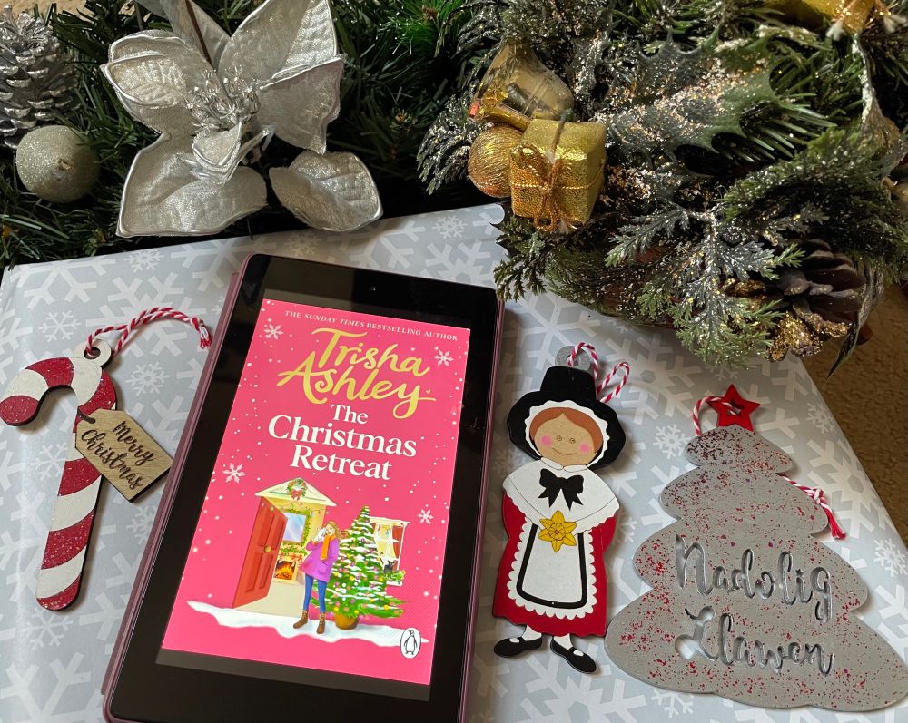 book cover of The Christmas Retreat by Trisha Ashley with tree decorations in the form of a striped red and white candy cane with the words Merry Christmas. A lady in a  Welsh costume and silver tree with the words Nadolig Llawen . With silver cone and flower and gold and green decorations above. 