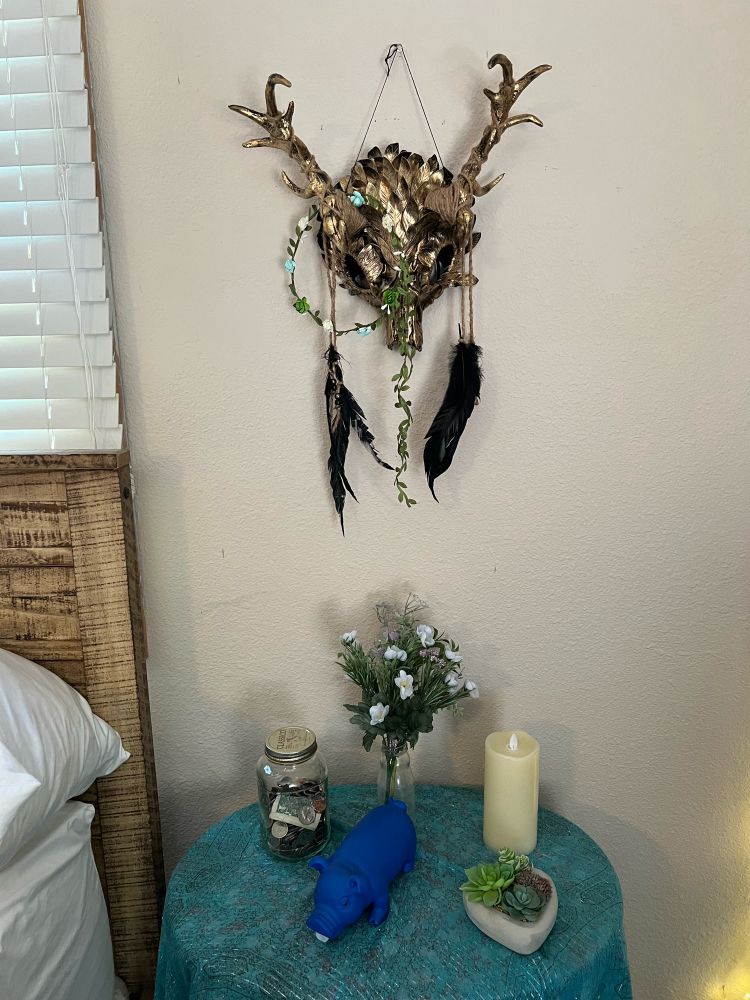 a gold antlered animal skull mask hanging on a beige wall with black feathers dangling from it and a flower crown perched on one of the antlers. below the mask is a small table with turquoise covering on it. on the table are a jar of coins, a fake succulent arrangement, some fake light blue flowers in a bottle, and a blue silicone pig (the pig makes little pig noises when you squeeze it and it brings me joy, thanks marj 💟)