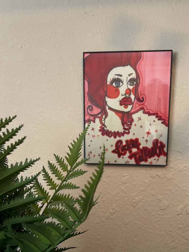 a self portrait of @rockettalksalot on tiktok where they drew themselves as a clown with a tall red bouffant. the drawing is framed and mounted on an otherwise blank piece of beige wall. a little fern from my desk is peeking into the frame of the photo.