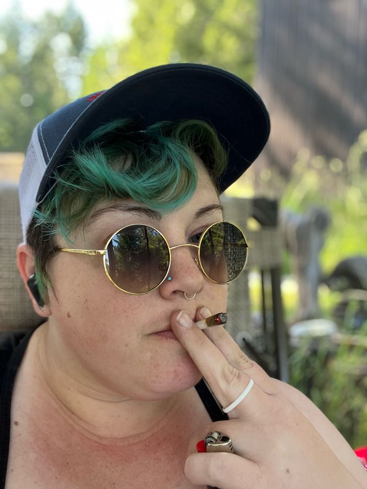 my face, smoking a joint, with short wavy green hair under a trucker hat, gold-rimmed round sunglasses, and a white aromantic ring on my left middle finger. i have a turquoise nose stud and a silver septum ring.