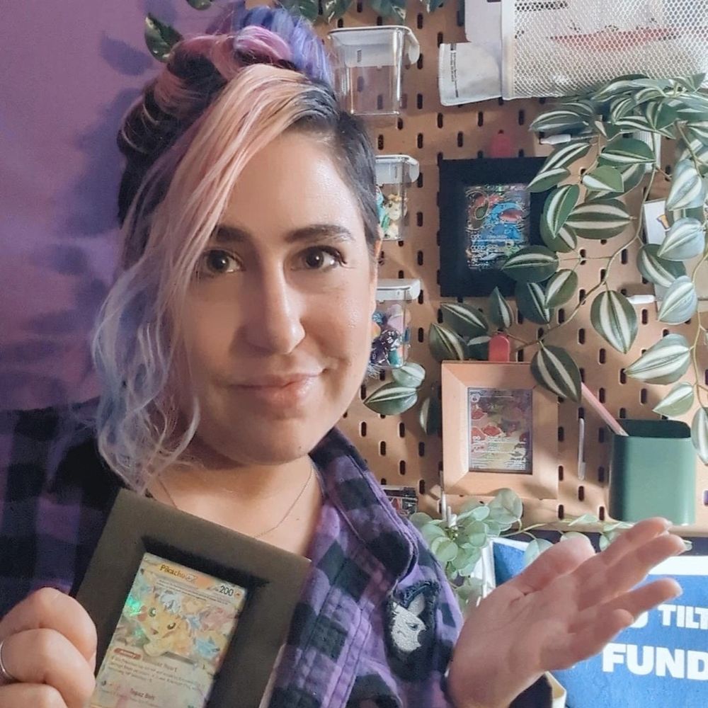 Nonbinary streamer FaunAndGames with pink, blue, and dark brown hair is wearing a purple and black checkered shirt. They are holding up a fancy Pikachu Pokémon card in a nice black frame towards the camera and smiling. They are showing off a plant covered board behind them with more Pokemon TCG cards in frames.